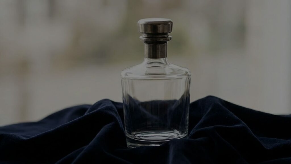 bespoke glass spirit bottle with a unique, heavy metallic closure, resting gracefully on a draped, dark navy velvet cloth in soft, natural daylight.